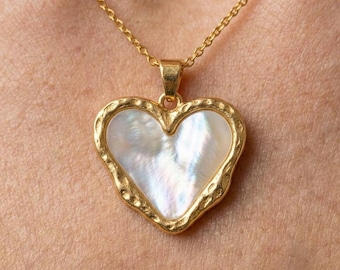 May include: A gold-toned necklace featuring a heart-shaped pendant with a white iridescent mother-of-pearl inlay. The pendant has a textured, hammered-style gold border and hangs from a delicate gold-toned chain link necklace.