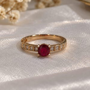 Op de afbeelding: Een gouden ring met een opvallende, rondgeslepen dieprode edelsteen in het midden, geflankeerd door een reeks kleinere, heldere, rondgeslepen stenen langs de band. De metalen ring heeft een gestructureerd, gesegmenteerd ontwerp met een glans.