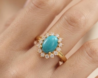 May include: A gold ring featuring a central oval turquoise gemstone surrounded by a halo of small, clear, round-cut white stones. The band is smooth and gold-toned, providing a classic setting for the vibrant blue-green turquoise center stone.