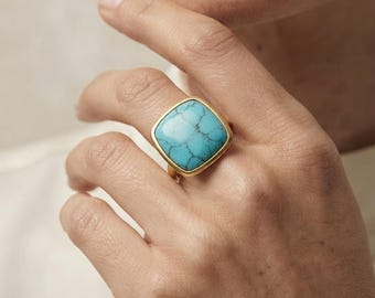 May include: A gold ring featuring a large, square-shaped turquoise stone with dark brown veining. The stone is set in a smooth, polished gold-toned metal bezel that frames the vibrant blue surface, creating a bold and prominent piece of jewelry.