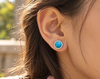 May include: A round stud earring featuring a smooth, vibrant turquoise cabochon center stone surrounded by a halo of small, clear, sparkling white crystals set in a polished silver-toned metal frame, worn on the earlobe.