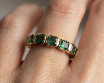 May include: A gold band ring featuring a series of emerald-cut green gemstones set in a continuous row. Each gemstone is encased in a gold bezel setting, creating a uniform and linked design that encircles the finger with vibrant green stones.
