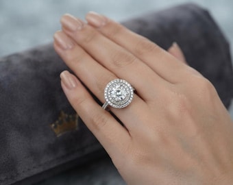 May include: A silver-toned ring featuring a large, round, brilliant-cut center stone surrounded by a double halo of smaller, round, clear stones. The band is thin and also encrusted with small, clear stones along the visible upper shank.