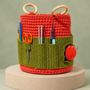 May include: A cylindrical crochet storage basket featuring a vibrant red upper section and a dark olive green lower section with multiple exterior pockets. The basket includes two light-colored wooden ring handles and holds various crochet hooks, orange scissors, and a round red pincushion with colorful pins.