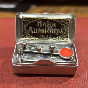 May include: A small silver-toned metal case containing a mechanical device with a bright orange circular accent. The interior lid features a black label with gold text reading Haka Autoknips Mod.I, set against a geometric gold-toned background pattern.