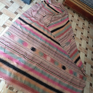 May include: A woven textile throw featuring horizontal stripes in shades of pink, peach, sage green, and black. The fabric displays a repeating pattern of small, dark, diamond-shaped motifs interspersed throughout the striped design, finished with a fringed edge.