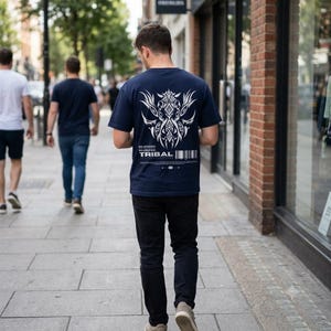 Puede incluir: Una camiseta azul marino de manga corta con un gran gráfico tribal blanco y simétrico en la espalda. Debajo del gráfico, el texto dice TRIBAL junto a un código de barras y texto más pequeño. La camiseta se combina con pantalones negros y zapatos claros.