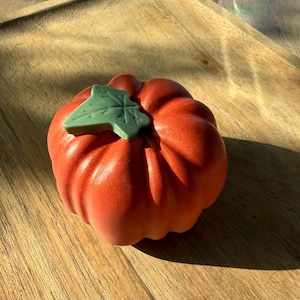 May include: A round, orange pumpkin-shaped object with a smooth, matte finish and deep vertical ridges. A single, flat, light green leaf-shaped accent is attached to the top center of the pumpkin, providing a contrasting color and texture.