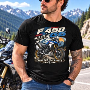 Könnte beinhalten: Ein schwarzes Kurzarm-T-Shirt mit einer Grafik eines blau-weißen Abenteuer-Motorrads. Der Text lautet F 450 GS NEXT GEN ADVENTURE und BUILT FOR WHAT'S AHEAD, ergänzt durch eine Berglandschaft, eine Sonne und ein Kompassrosen-Symbol.