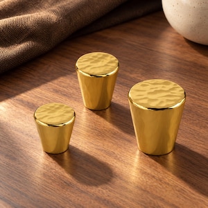 Puede incluir: Tres pomos de armario de color dorado con una base lisa y cónica y una superficie superior texturizada y martillada. Los pomos varían en tamaño y presentan un acabado metálico reflectante que capta la luz, creando sombras.
