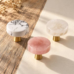 May include: Three round cabinet knobs with flat tops and brass-colored conical bases. One knob features a white surface with black veining, one is a translucent pale pink, and one is a semi-translucent off-white stone material.