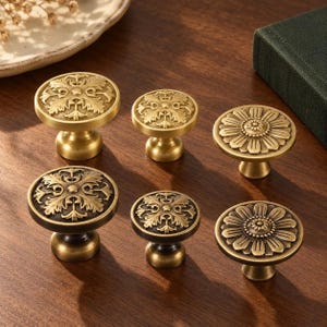 Puede incluir: Un conjunto de seis pomos redondos de metal para armarios con acabado en latón. Tres pomos presentan un patrón floral de hojas en relieve, mientras que los otros tres muestran un diseño de pétalos radiales. Los pomos varían en tamaño y tienen una pátina oscura.