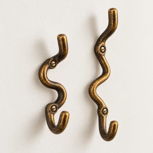 May include: Two wall-mounted hooks with a wavy, serpentine silhouette and a dark, antiqued brass finish. Each hook features two visible flat-head screws for mounting, with a curved hook at the bottom and a rounded top end for hanging items.