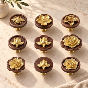 Puede incluir: Una colección de nueve pomos para armarios con bases de madera de color marrón oscuro, circulares o festoneadas, con detalles en metal dorado. Los diseños metálicos incluyen una abeja, varias flores y capullos cerrados.