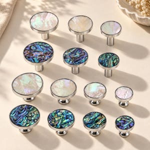 Puede incluir: Una colección de doce pomos circulares para armarios dispuestos en cuatro filas. Cada pomo cuenta con una base de metal plateado y una incrustación redonda de nácar blanco iridiscente o de concha de abulón con tonos azules y verdes.