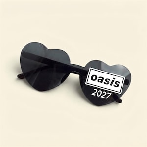 May include: Heart-shaped sunglasses with black lenses and black frames. The right lens features a white rectangular graphic containing the black text "oasis" with the white text "2027" positioned directly underneath the rectangle.