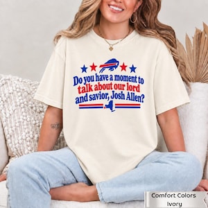 Op de afbeelding: Een ivoorkleurig T-shirt met korte mouwen en rode en blauwe tekst: "Do you have a moment to talk about our lord and savior, Josh Allen?" Het ontwerp bevat een blauw buffelsilhouet, rode en blauwe sterren en een blauwe kaart van New York.