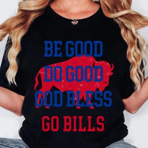 May include: A black short-sleeved t-shirt featuring a large red silhouette of a buffalo. Overlaid on the buffalo is the blue text "BE GOOD DO GOOD GOD BLESS GO BILLS" in a bold, block-style font. The design has a slightly distressed texture.
