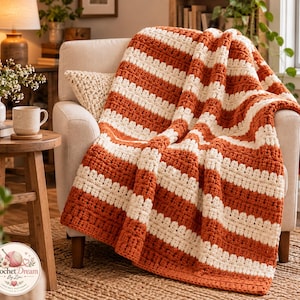 Può includere: Una coperta all'uncinetto con strisce orizzontali alternate di filato color arancio bruciato e crema. Il tessuto presenta un motivo a punti in rilievo su tutta la superficie, creando un aspetto spesso e morbido mentre è drappeggiato su una poltrona neutra.