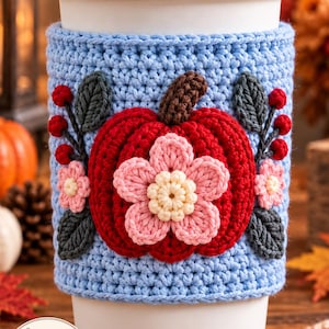 May include: A light blue crochet cup sleeve featuring a central red pumpkin with a brown stem and a layered pink and cream flower. The design includes dark grey leaves and small red berries on both sides. A logo reads Crochet Dream By Zin.