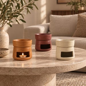 May include: Three ceramic wax melt warmers in terracotta, dark red, and cream colours. Each warmer features a cylindrical base with a rectangular cutout for a tea light candle and a bowl on top. One warmer contains a lit tea light candle.