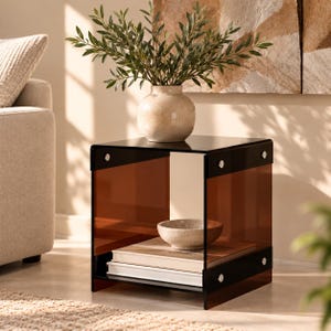 Peut inclure: Une table d'appoint carrée composée de panneaux en acrylique ambré translucide et de supports en acrylique noir, avec des fixations argentées. La table comprend une étagère inférieure supportant deux livres empilés et un bol en pierre.