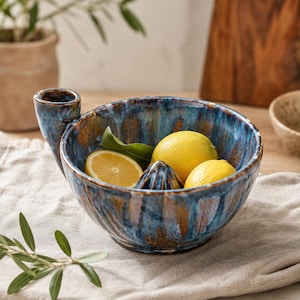 Może przedstawiać: Ceramiczna wyciskarka do cytrusów z wykończeniem z reaktywnego szkliwa w kolorze głębokiego błękitu i brązu. Misa posiada centralny stożek do wyciskania soku oraz boczny uchwyt do nalewania. Wewnątrz znajdują się dwie całe cytryny i połówka cytryny.