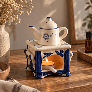 Peut inclure: Un brûle-parfum en céramique composé d'une base carrée ornée de motifs floraux bleus et blancs et d'un sommet en forme de théière. La base présente une arche ouverte contenant une bougie chauffe-plat allumée dans un petit support blanc.