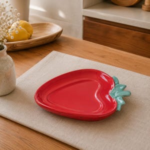 Peut inclure: Un plat de service en céramique brillante en forme de fraise, doté d'un corps rouge vif et d'une tige vert clair sur le dessus. Le plat présente une surface lisse et réfléchissante ainsi qu'une silhouette en forme de cœur pour les aliments.