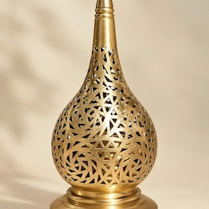 Op de afbeelding: Een goudkleurige metalen lamp met een druppelvormig lichaam voorzien van een ingewikkeld, lasergesneden geometrisch patroon van overlappende lijnen. De lamp heeft een puntige knop aan de bovenkant en staat op een gladde, ronde voet.