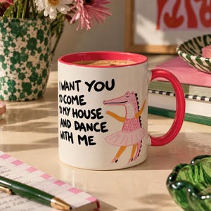 Peut inclure: Un mug en céramique blanche avec un intérieur et une anse rose vif. L'extérieur présente une illustration de crocodile rose portant un tutu et des chaussons de danse, accompagnée du texte noir : I WANT YOU TO COME TO MY HOUSE AND DANCE WITH ME.