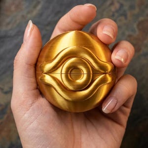 May include: A spherical gold-coloured object featuring a raised, stylised eye design on its surface. The object is constructed with a horizontal seam across the middle, indicating two halves that fit together to form a complete, rounded shape.