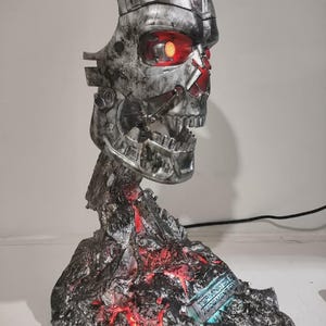 May include: A metallic silver robotic skull sculpture with a glowing red eye, mounted on a jagged, dark base with integrated red lighting effects. The base features a small plaque with the text Terminator 2 Judgment Day in blue lettering.