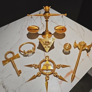 May include: A collection of gold-colored props including a balance scale, an ankh key, a circular bracelet, a triangular pyramid with an eye symbol, a spherical object, a multi-pointed star emblem, and a long-handled axe with a central eye motif.