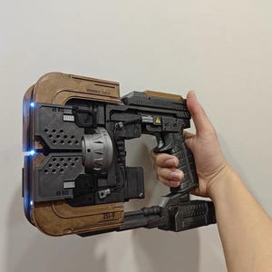 Puede incluir: Un accesorio de cortador de plasma de ciencia ficción con un acabado marrón desgastado y gris oscuro. El dispositivo tiene un marco frontal rectangular con luces LED azules, un emisor circular central y un mango texturizado. Incluye las marcas 211-V y XS 972.