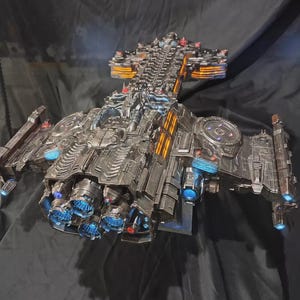 Könnte beinhalten: Ein detailliertes Modell eines metallisch grauen Raumschiffs mit einem komplexen Rumpf, zahlreichen mechanischen Komponenten, mehreren hinteren Triebwerken mit blauem Leuchten und Seitenpaneelen mit leuchtend orangefarbenen Streifen.
