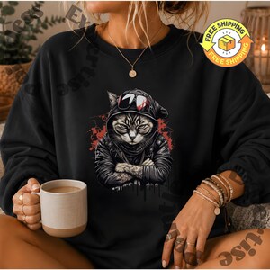 May include: A black crewneck sweatshirt featuring a graphic print of a cat wearing a black hooded jacket and sunglasses with its arms crossed. The cat illustration is set against a red paint splatter background. The sweatshirt has long sleeves.