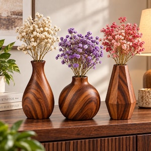 Op de afbeelding: Drie houten vazen in verschillende vormen met droogbloemen. De linker vaas is druppelvormig met witte bloemen, de middelste is bolvormig met paarse bloemen en de rechter is gefacetteerd met roze bloemen. Alle vazen hebben een donkere houtnerf.