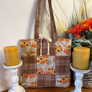 May include: A patchwork tote bag featuring a repeating pattern of pumpkins, sunflowers, and autumn leaves, alternating with brown and white plaid fabric panels. The bag includes two long shoulder straps and a white drawstring tie at the top center.