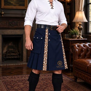 May include: A navy blue kilt featuring a leopard print panel, a large cargo pocket with silver-toned buttons, and a black leather belt. The kilt displays the Heritage Mantle Craft logo in gold embroidery and is paired with black knee-high socks and boots.