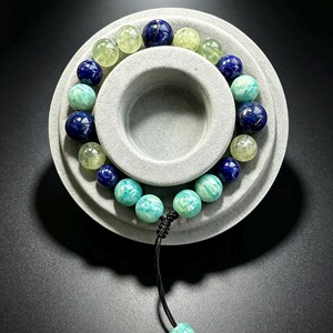 May include: A beaded bracelet featuring alternating round beads in deep blue, pale green, and turquoise hues. The bracelet is strung on a black cord with an adjustable sliding knot closure, finished with a single turquoise bead at the end.