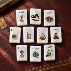 Puede incluir: Diez fichas rectangulares blancas con acabado nacarado, cada una con ilustraciones únicas de gatos y arreglos florales. Las fichas muestran varios números, la palabra JOKER y la palabra FLOWER, con detalladas ilustraciones de gatos.