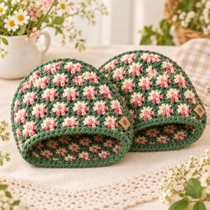 Könnte beinhalten: Zwei gehäkelte Mützen mit einem wiederkehrenden Blumenmuster in Dunkelgrün, Creme und Rosa. Jede Mütze hat einen einfarbigen, dunkelgrünen Rippsaum und ein kleines, quadratisches, hellbraunes Etikett in Lederoptik mit einem Blumensymbol.