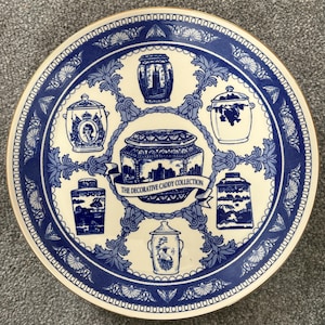 May include: A round ceramic plate with a cream-colored center and a wide blue border featuring intricate patterns. The center displays six illustrations of various caddy jars, with a central banner reading THE DECORATIVE CADDY COLLECTION.