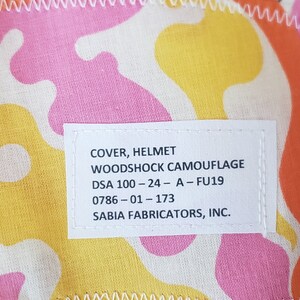 May include: A white label with black text that reads "COVER, HELMET WOODSHOCK CAMOUFLAGE DSA 100-24-A-FU19 0786-01-173 SABIA FABRICATORS, INC."