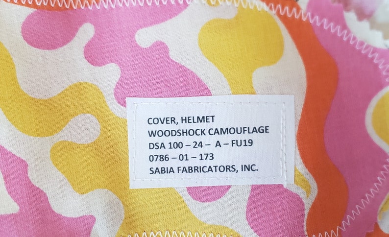 May include: A white label with black text that reads "COVER, HELMET WOODSHOCK CAMOUFLAGE DSA 100-24-A-FU19 0786-01-173 SABIA FABRICATORS, INC."