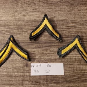 May include: Three yellow and black Private E2 rank patches arranged on a wooden surface. The patches are chevron-shaped with a yellow center and black border. A handwritten note is also present.