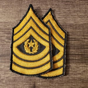 May include: Two embroidered military rank insignia. Each patch features a gold star and wreath emblem on a black background, with gold chevrons on a black base. The edges are outlined with a black border. The patches are likely for a Sergeant First Class.