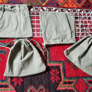May include: Four olive green drawstring bags of varying sizes are arranged on a patterned red rug. The bags appear to be made of a durable fabric. A clear ruler with white markings is also visible.
