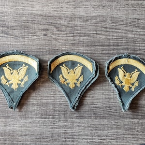May include: Three vintage military patches with a blue-green background and gold embroidered details. Each patch features a gold eagle emblem and a gold arched top. The patches are arranged on a wooden surface.